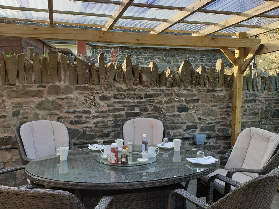 A covered outdoor dining area with a stone wall backdrop, wicker chairs, and a glass table set for four.