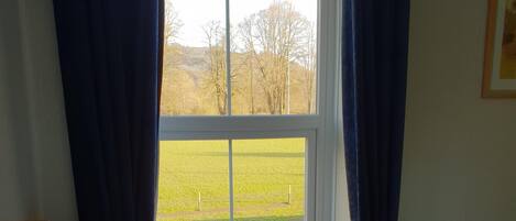 A bedroom with a bed, a window with curtains, a view of a green field, and a lamp on a bedside table.