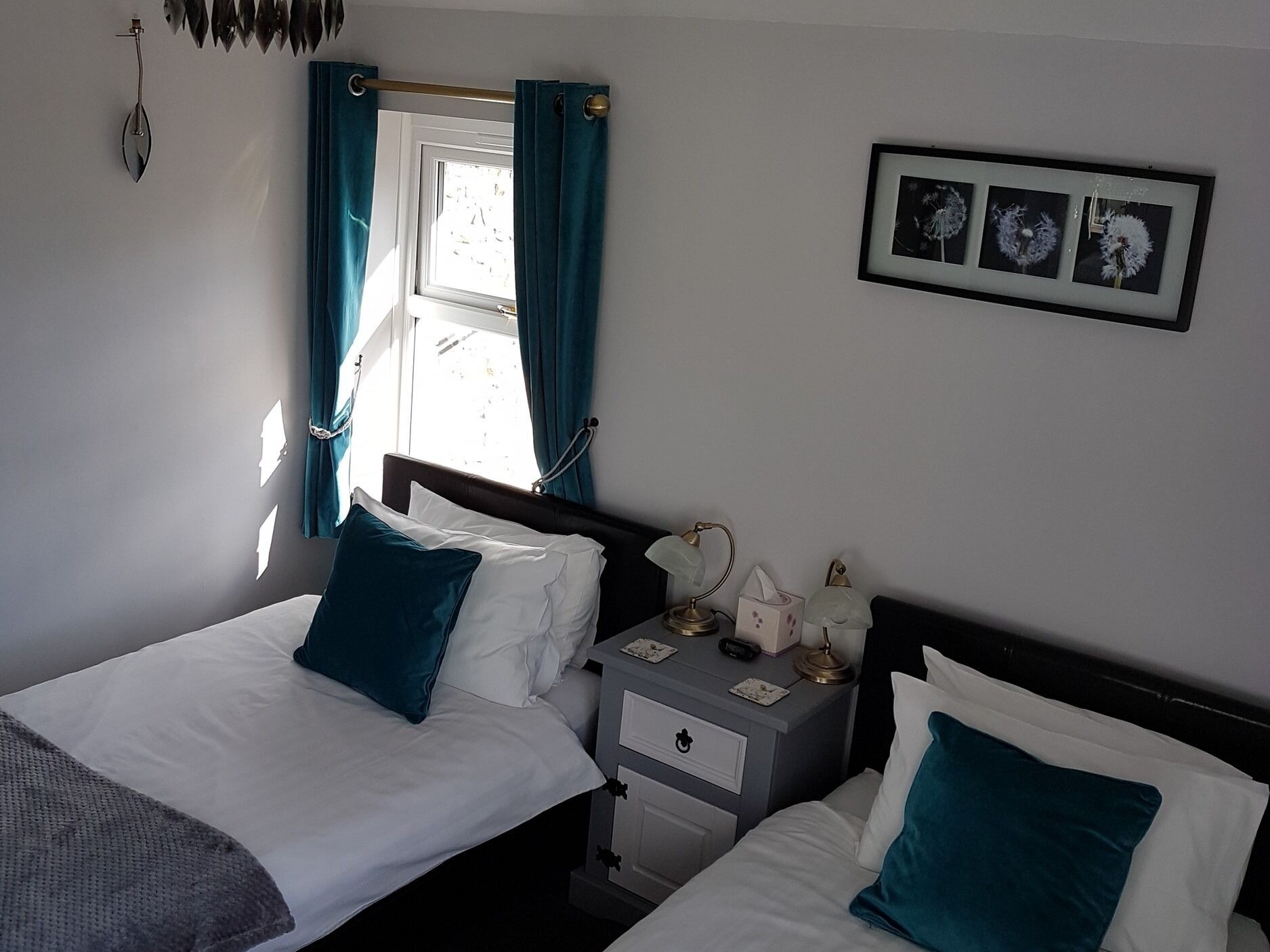 Une chambre avec deux lits, une table de chevet, une commode, une fenêtre avec des rideaux et un tableau encadré au mur.