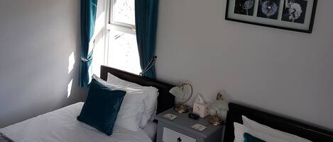 Une chambre avec deux lits, une table de chevet, une commode, une fenêtre avec des rideaux et un tableau encadré au mur.