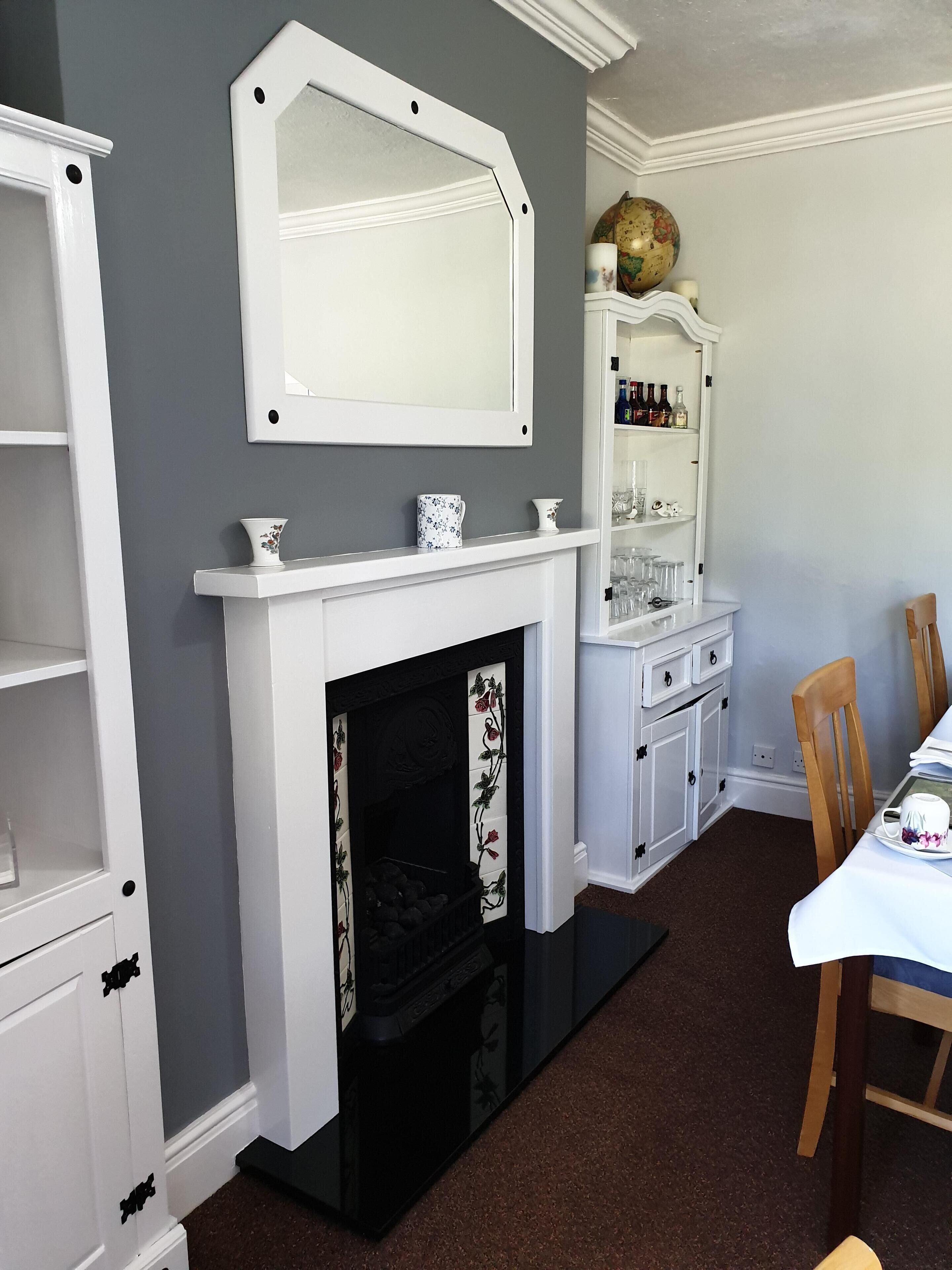 A room with a white fireplace, a mirror above it, a shelf with bottles, and a dining table with chairs.
