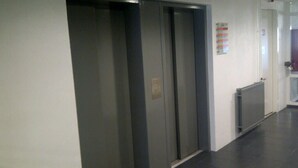 Un pasillo con tres puertas de ascensor cerradas, un radiador y una puerta con un cartel en la pared.