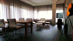 Una habitación de hotel moderna con una mesa de comedor de madera, sillas de oficina, un sofá, un televisor sobre un mueble bajo y ventanas amplias con cortinas.