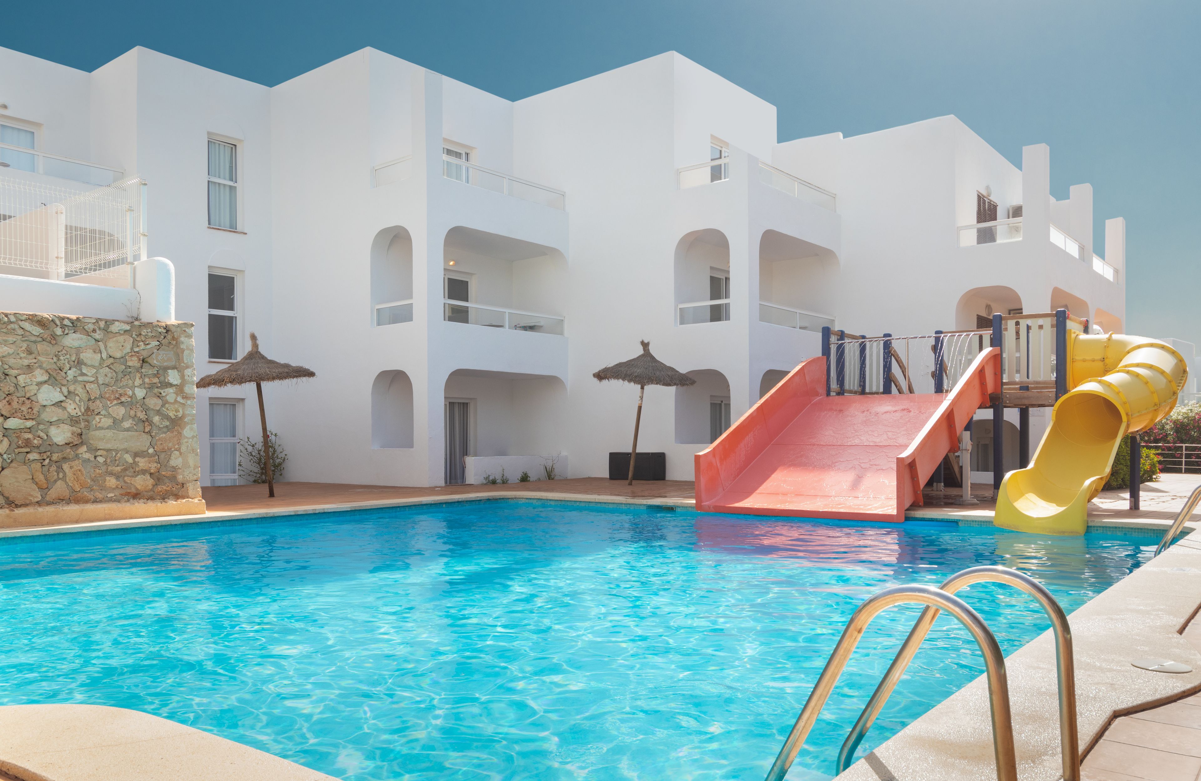 Una piscina con un tobogán rojo y amarillo, rodeada de edificios blancos con balcones y un muro de piedra.