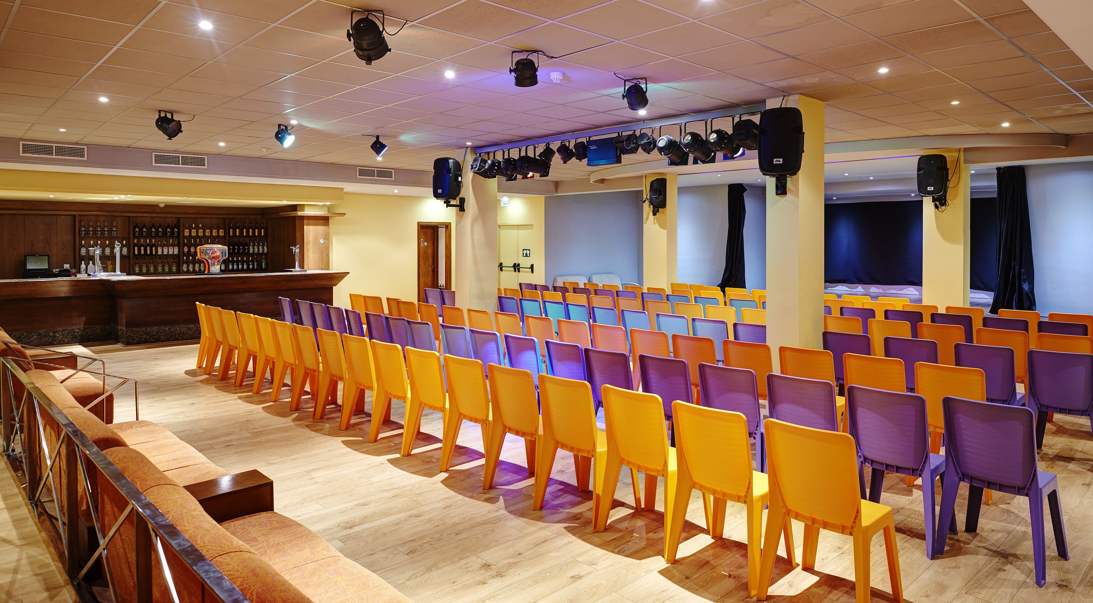 A conference room with rows of chairs, a stage with lighting equipment, and a bar area with bottles.