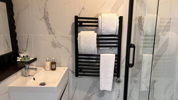 A bathroom with a marble-tiled wall, a white sink, a black towel rack with white towels, and a glass shower enclosure.