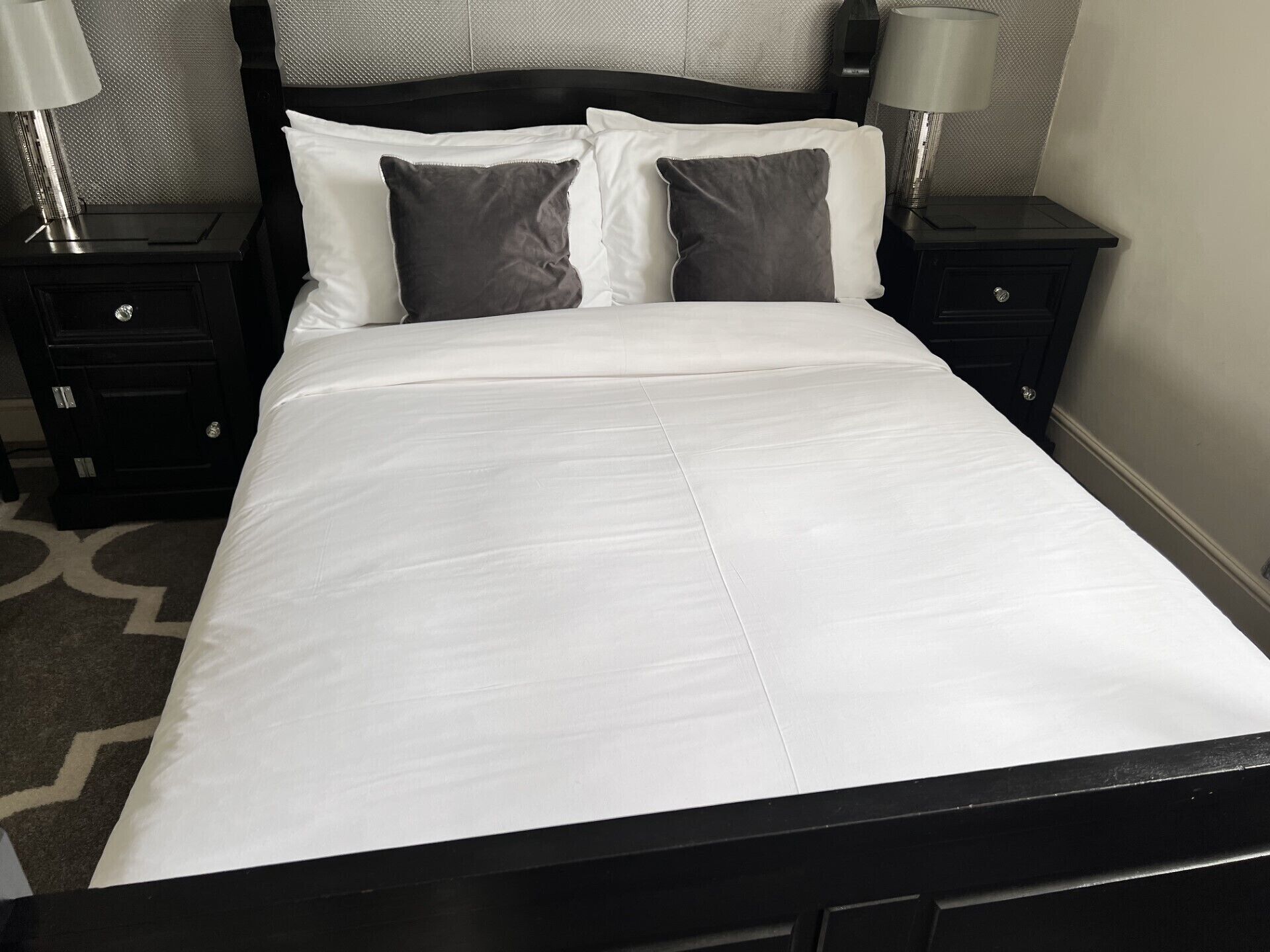 Een net opgemaakt bed met witte lakens en twee donkere kussens, met aan beide zijden nachtkastjes met lampen, in een kamer met een kamerbreed tapijt.