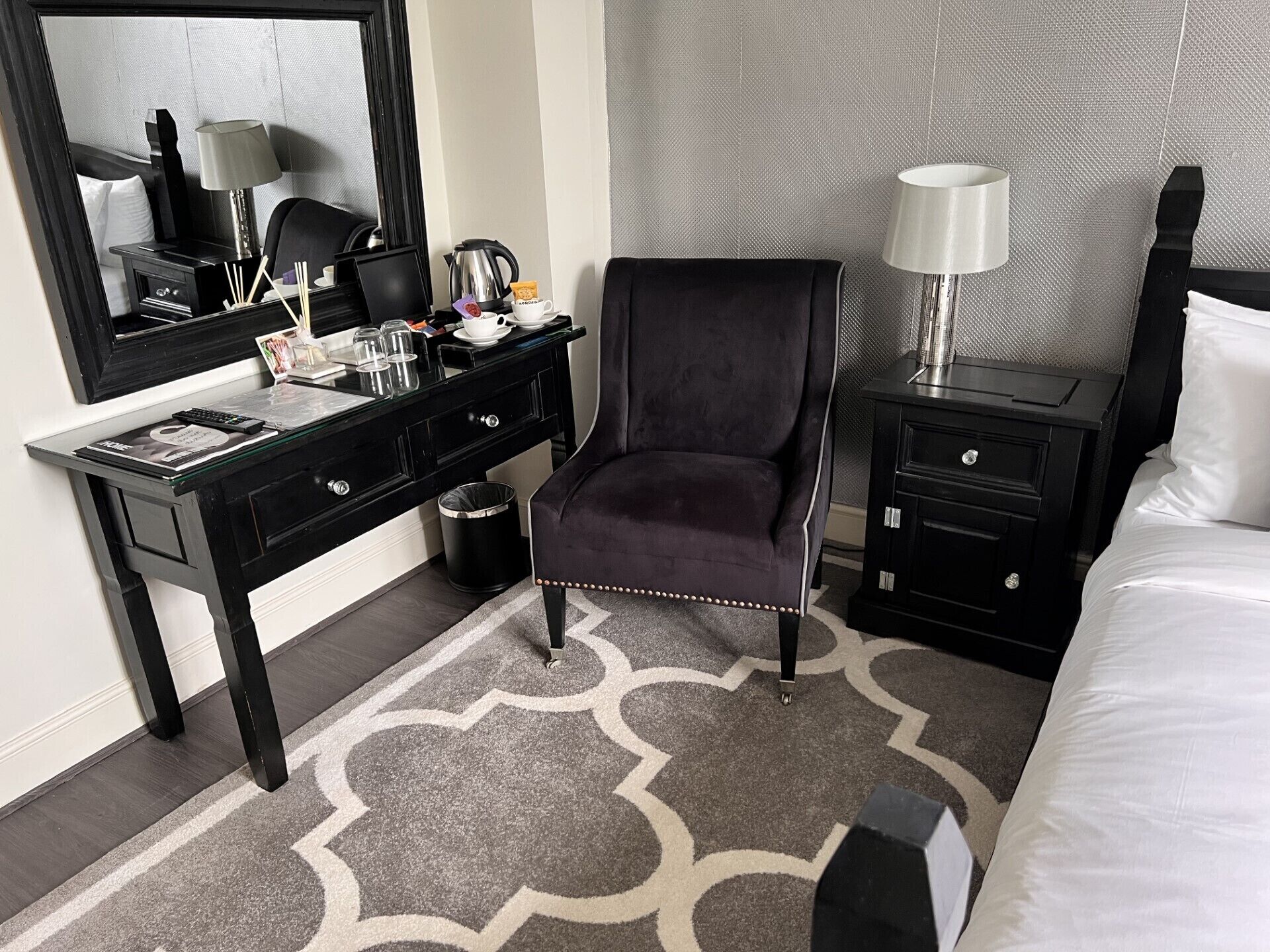 Habitación de hotel con cama, escritorio con computadora, silla, mesita de noche, lámpara y cesto de basura.