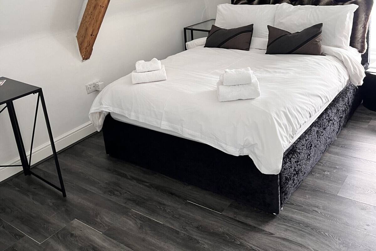 A modern bedroom with a sloped ceiling, a dark headboard, a bed with white linens and multiple pillows, a small bedside table, and a hanging light fixture.