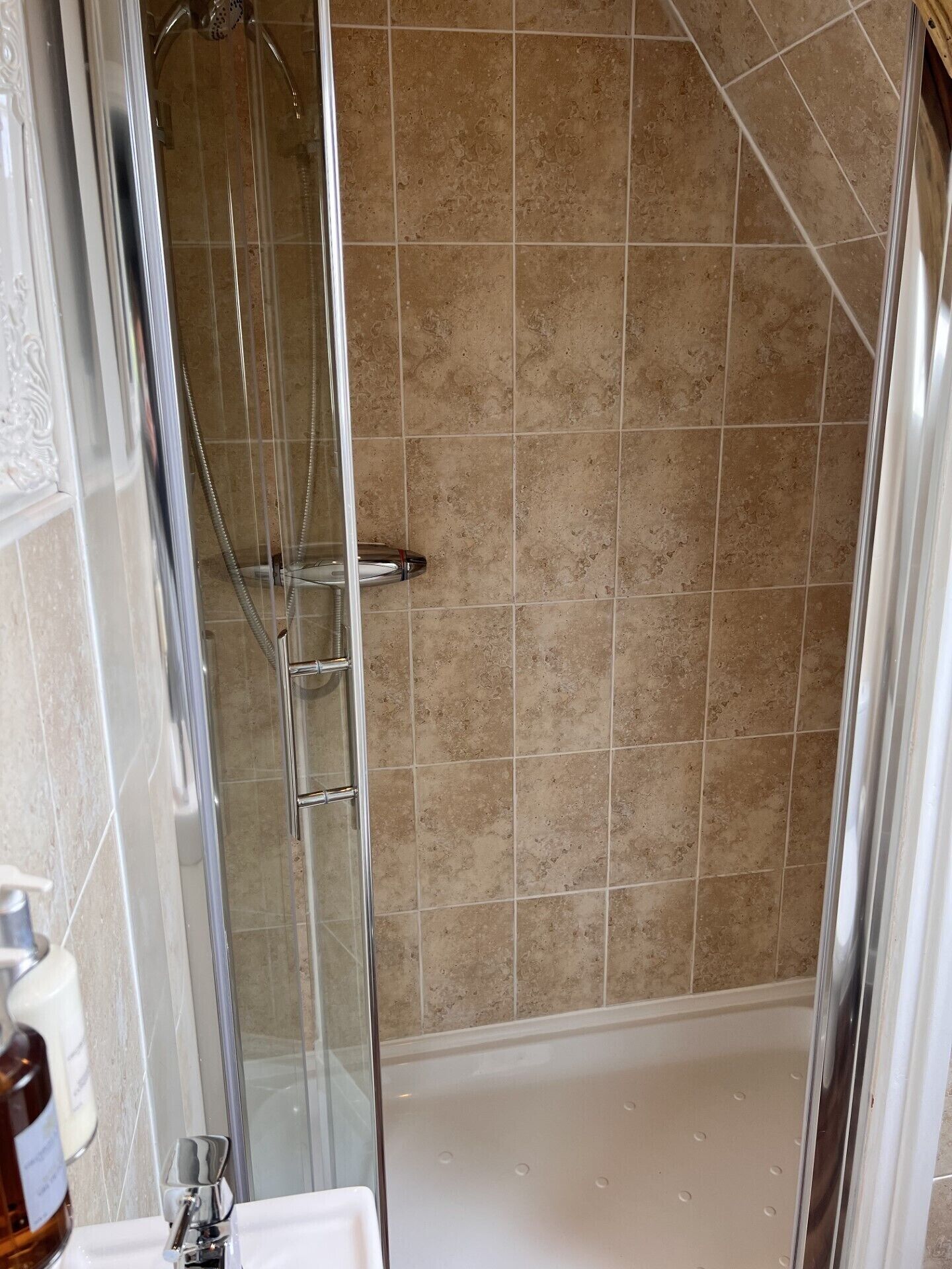 Una ducha con cerramiento de vidrio, azulejos color beige, un cabezal de ducha cromado y una bañera blanca.