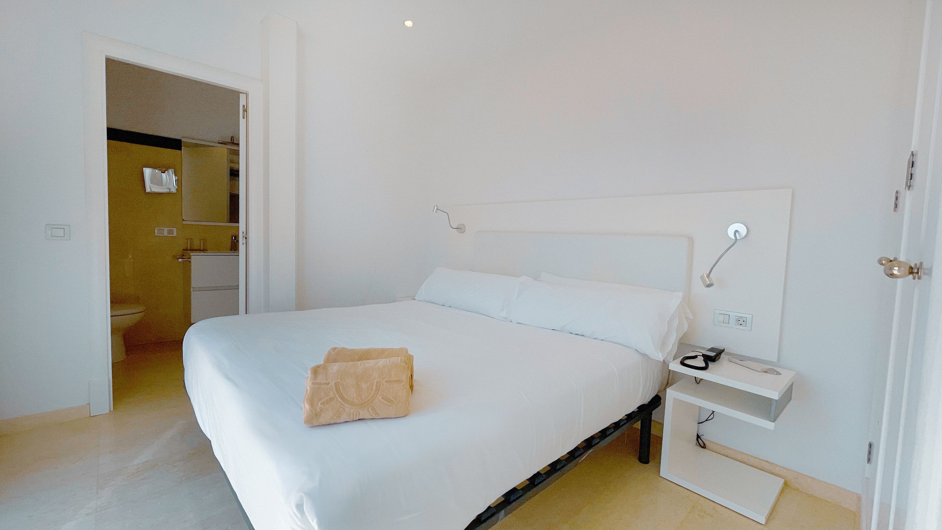 A neatly made bed with a white comforter and pillows, a bedside table with a lamp, and a small shelf.