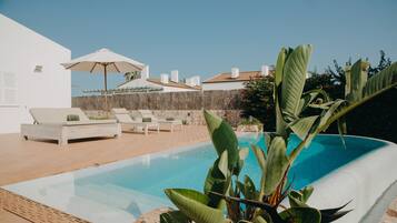 A swimming pool with lounge chairs and an umbrella, surrounded by plants and a stone wall.