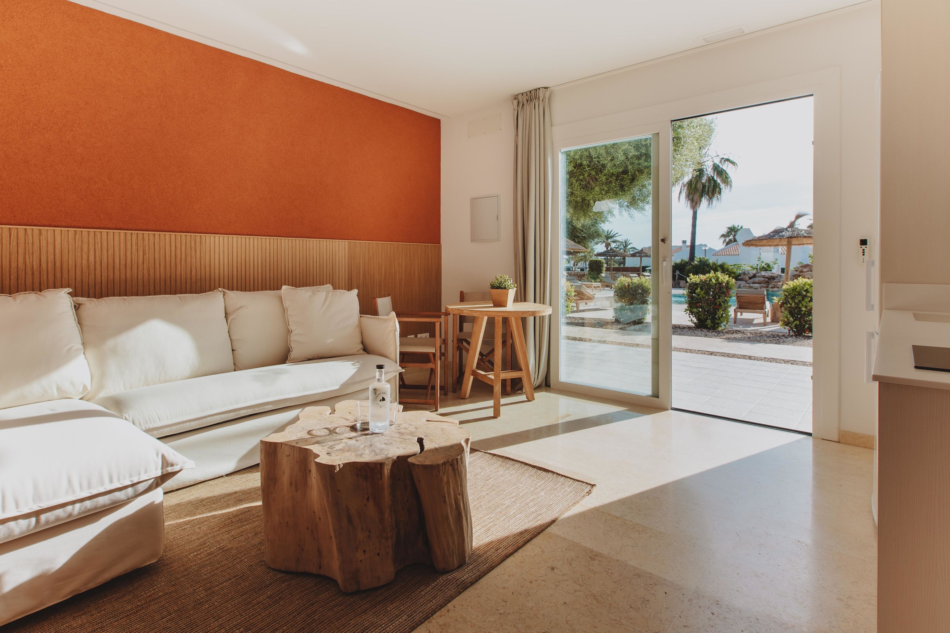 A modern hotel room with a large sofa, a wooden coffee table, and a view of the outdoor pool area.