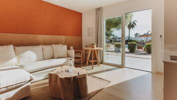 A modern hotel room with a large sofa, a wooden coffee table, and a view of the outdoor pool area.