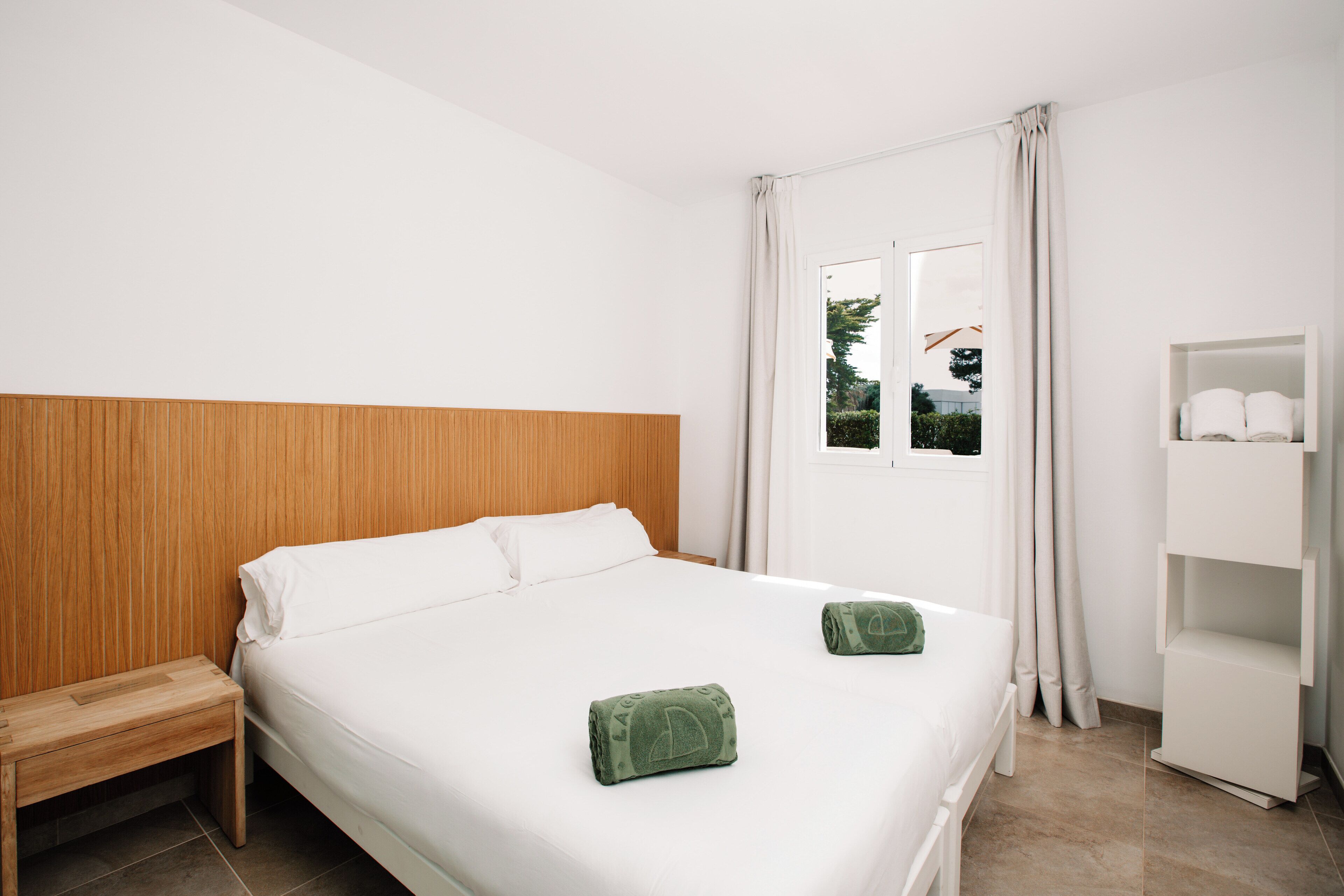 Une chambre d’hôtel avec une tête de lit en bois, un grand lit avec du linge de lit blanc, deux serviettes vertes, une table de chevet en bois, une étagère blanche et une fenêtre avec des rideaux.