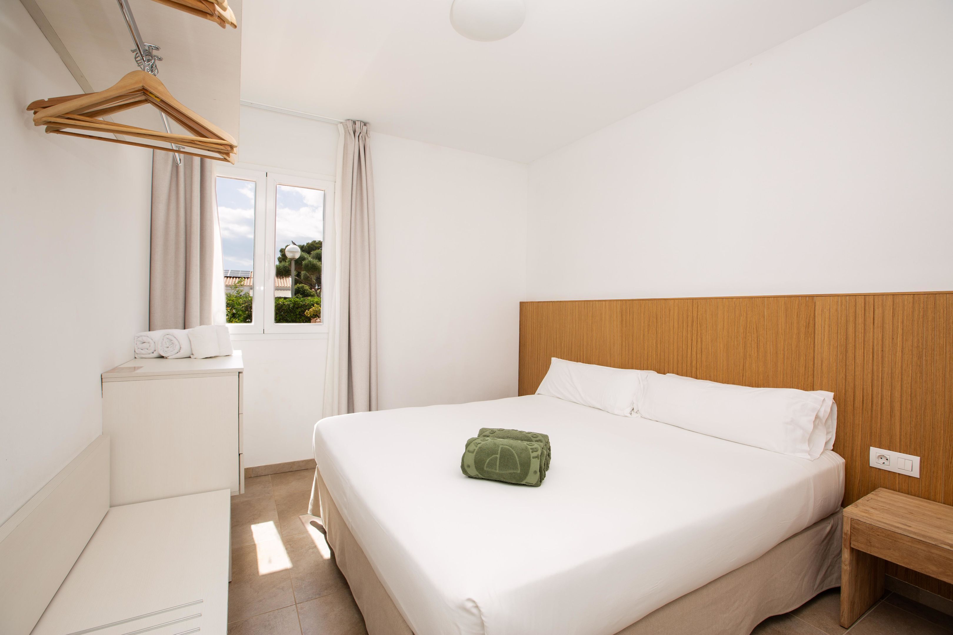 A hotel room with a bed, a wooden headboard, a bedside table, a hanging wooden hanger, and a window with curtains.