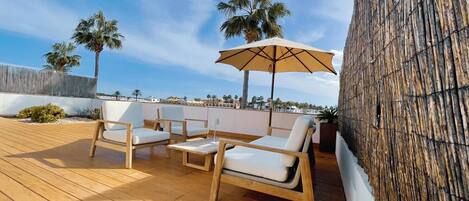 Una terraza de madera con mobiliario exterior y un gran paraguas, con vistas a una zona costera con palmeras y cielo despejado.