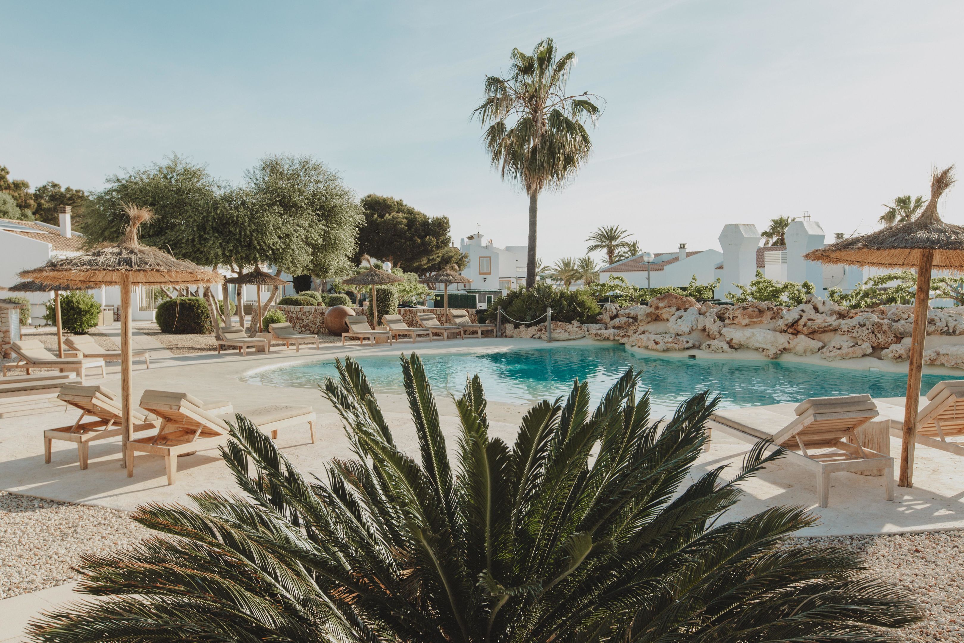 A pool area with lounge chairs and umbrellas, surrounded by palm trees and a rocky landscape.