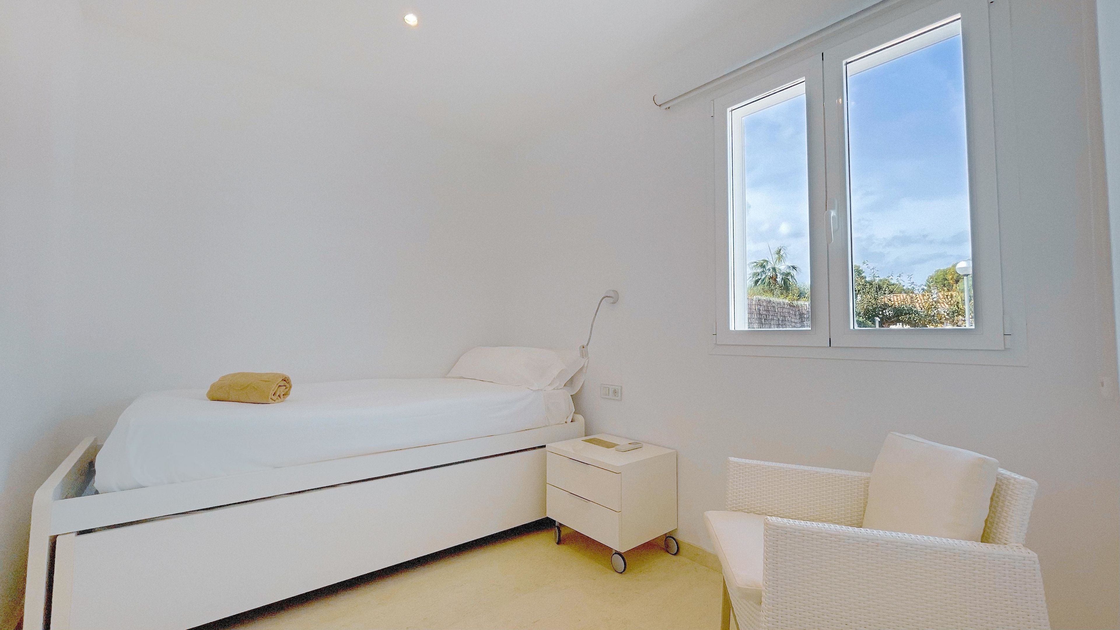 Un dormitorio con cama, mesita de noche, silla y ventana con vista a palmeras.