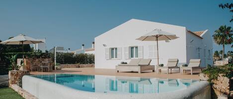 Una villa moderna y blanca con piscina, mobiliario exterior y un cielo azul despejado.