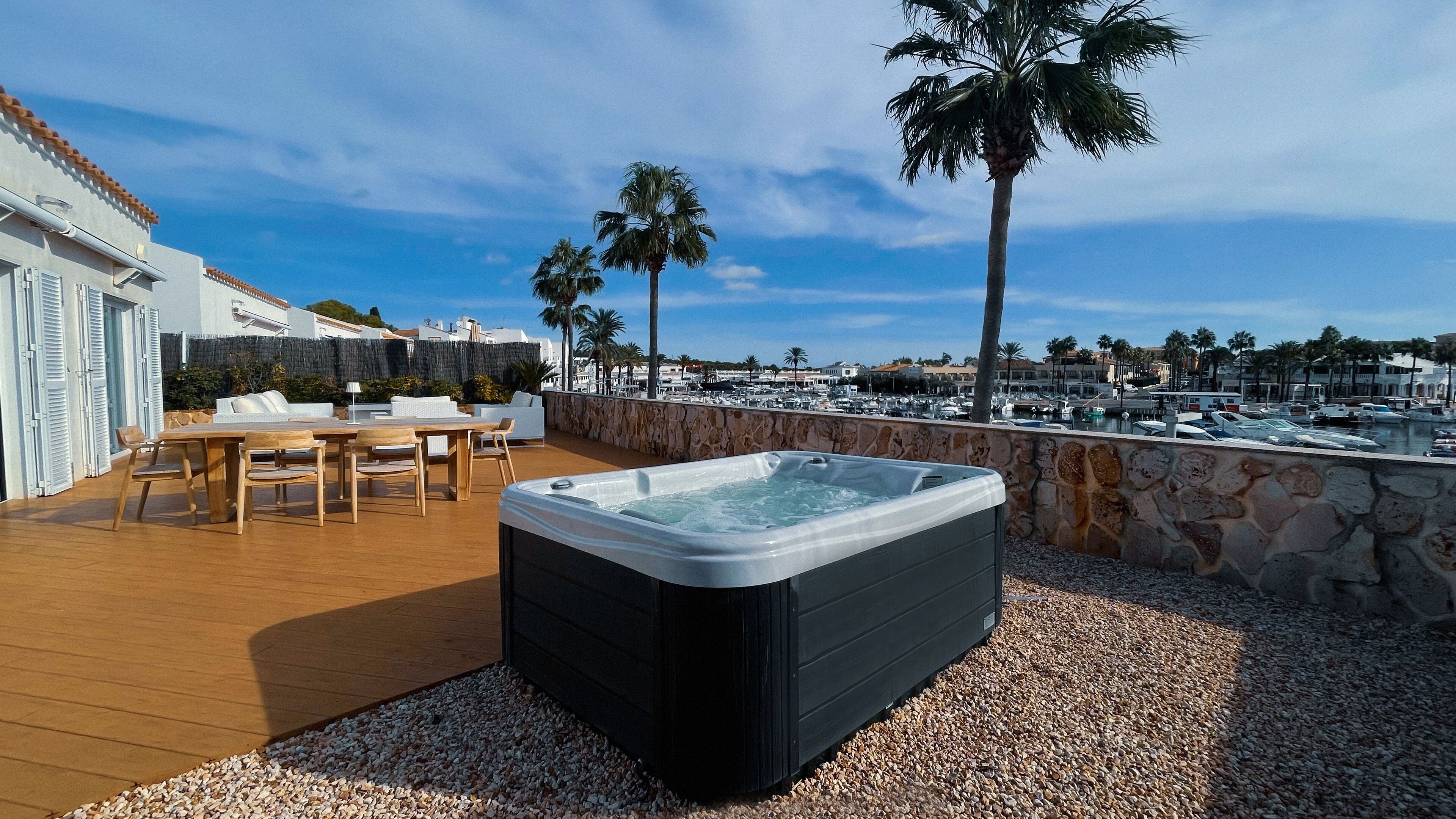 A rooftop terrace with a hot tub, wooden furniture, and a stone wall overlooking a marina with boats and palm trees.