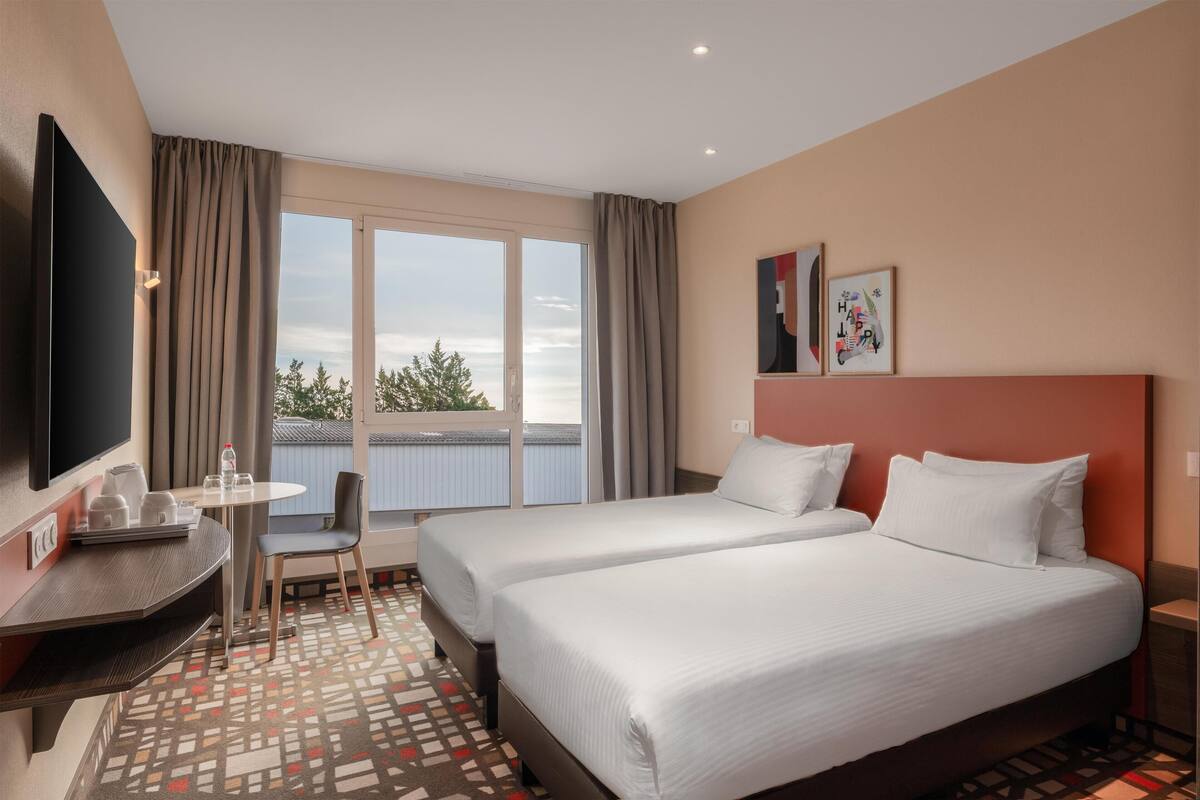 A hotel room with two beds, a desk with a chair, a TV, and a window with curtains.