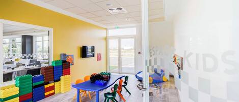 Un área de juegos infantil, de colores brillantes, con una mesa, sillas, un televisor y una ventana con vistas al exterior.
