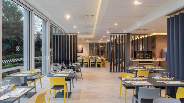 A modern restaurant with a long dining table, yellow chairs, and a view of trees through large windows. - Spark by Hilton Lyon Eurexpo (Genas)