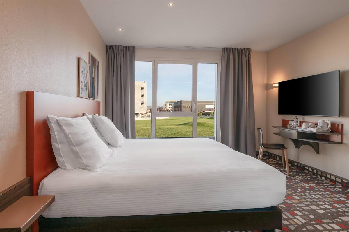A hotel room with a large bed, a TV mounted on the wall, a desk with a chair, and a view of the outside.
