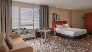 A hotel room with a bed, a sofa, a small table with chairs, and a large window offering a view of buildings and trees. - Spark by Hilton Lyon Eurexpo (Genas)