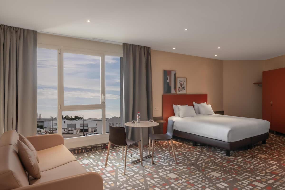 A hotel room with a bed, a sofa, a small table with chairs, and a large window offering a view of buildings and trees.
