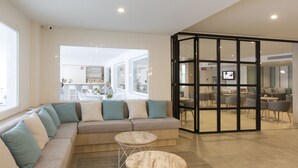 A modern hotel lobby with a large sectional sofa, two round tables, and a glass partition separating the seating area from the dining area. - Hotel HSM Linda Playa (Calvia)