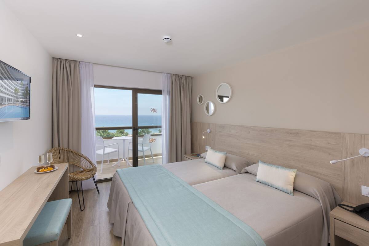 A hotel room with two beds, a desk with a chair, a balcony with a view of the sea, and a mounted TV.