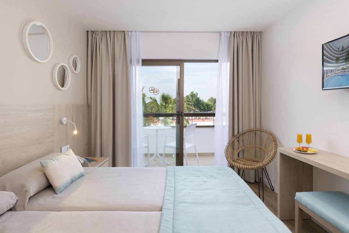 A hotel room with two beds, a desk with a chair, a balcony with a table and chairs, and a view of the outdoors.