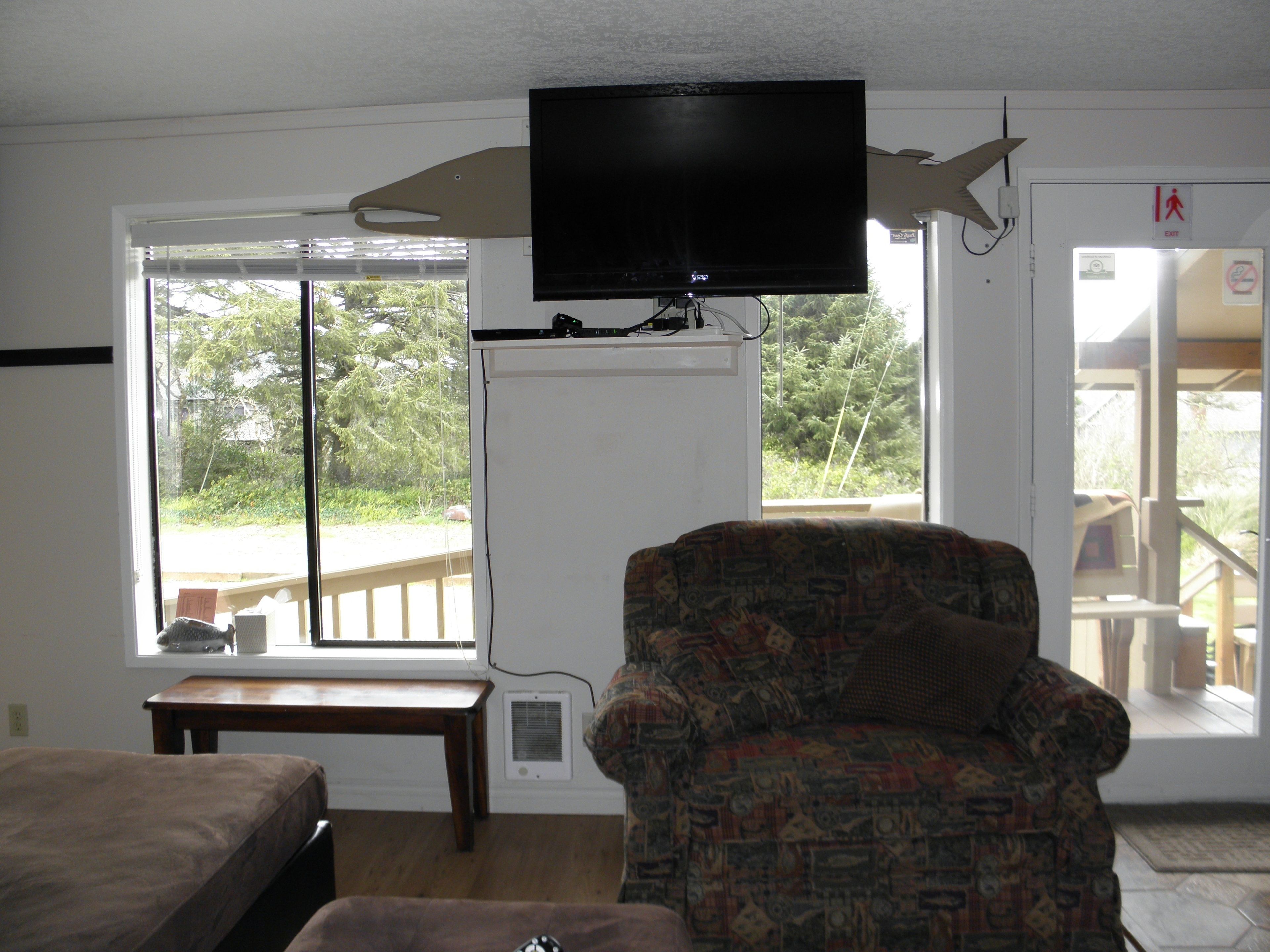 Una habitación con un televisor montado en la pared, un sillón estampado, una mesa de madera y una ventana con vistas a zonas verdes.