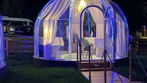 Uma estrutura em cúpula transparente com um deck de madeira que a leva até o interior, onde se encontram uma mesa e cadeiras.