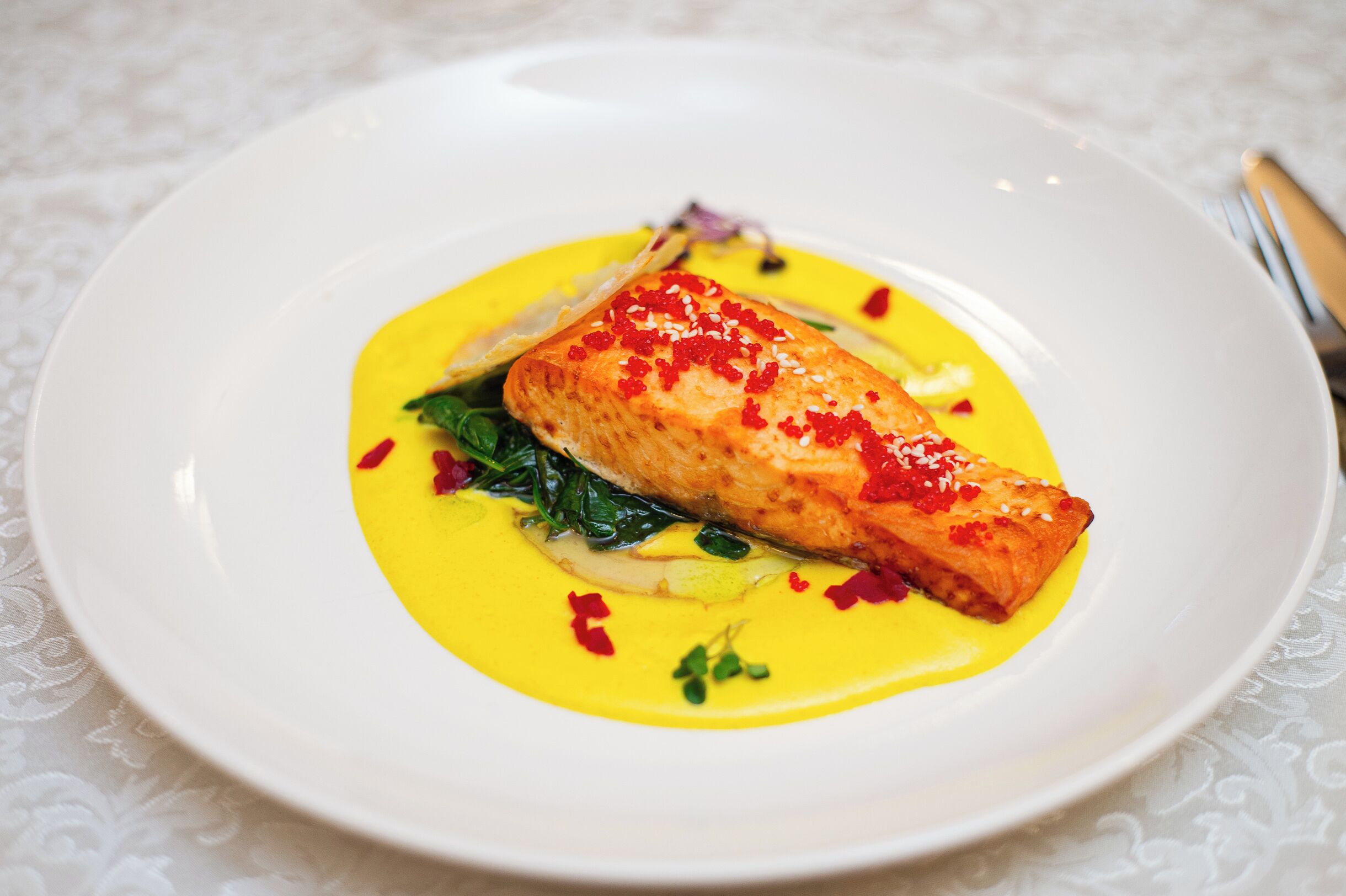 Un plato blanco con un trozo de salmón cubierto de caviar rojo, rodeado de salsa amarilla y adornado con vegetales verdes.