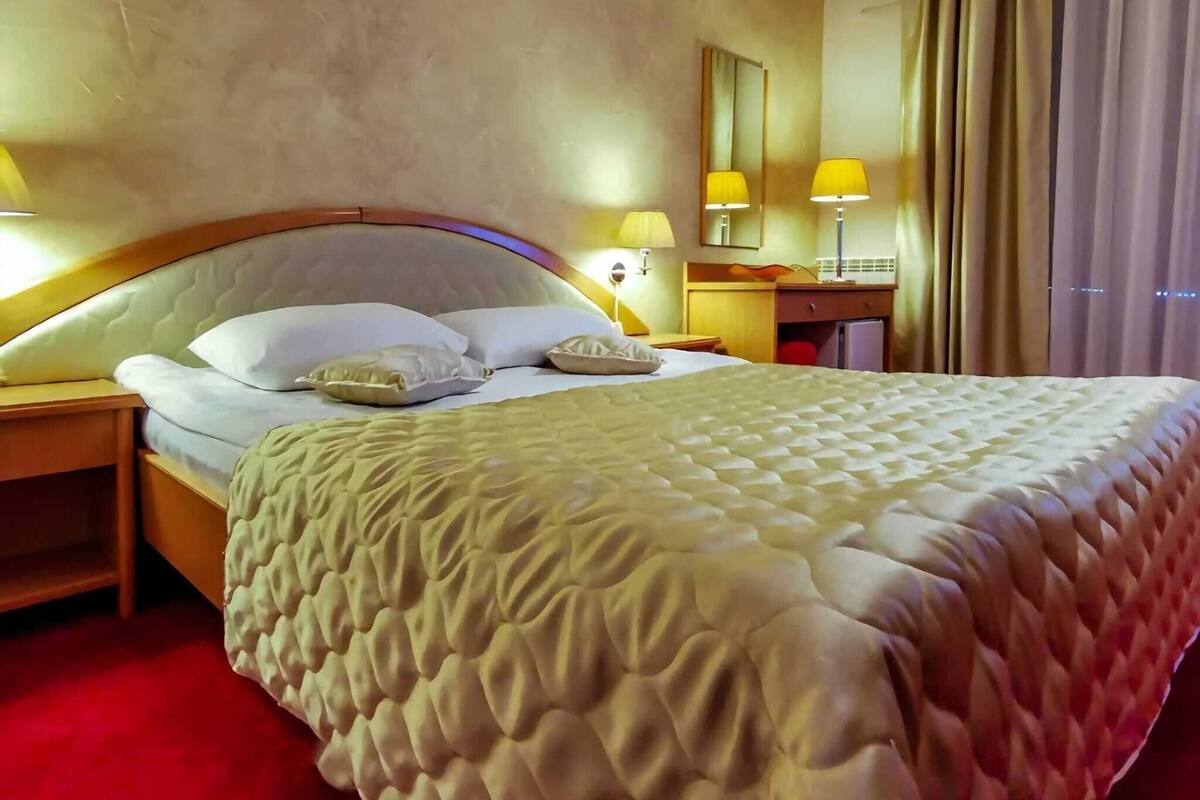 A hotel room with a large bed, two bedside lamps, a nightstand, a wooden headboard, a mirror, a desk with a lamp, and a window with curtains.