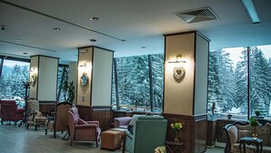 Um hall amplo com várias áreas de estar, grandes janelas com vista para árvores cobertas de neve e uma lareira com um espelho decorativo.