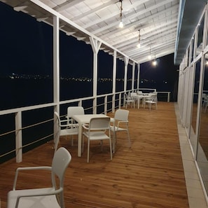 An outdoor covered area with wooden flooring, white chairs, and tables, overlooking a cityscape at night.