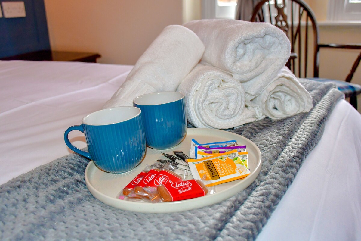 Ein ordentlich bezogenes Bett mit weißen Leinen, ein Tablett mit zwei blauen Tassen, zusammengerollte Handtücher und Frühstücksartikel, darunter Kekse und Tee.