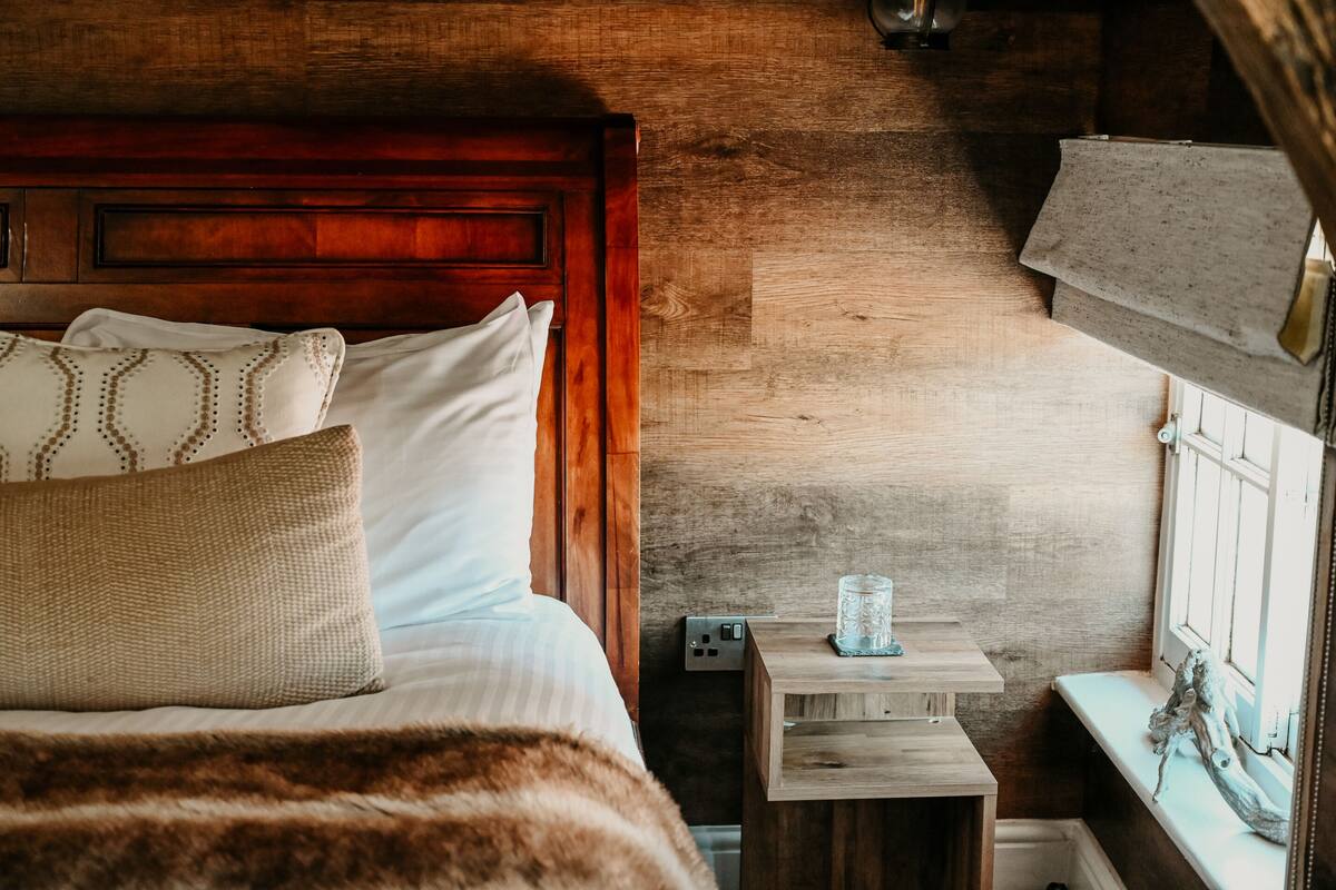 Una cama de madera con ropa de cama blanca y almohadas estampadas, una mesita de noche pequeña con un recipiente de vidrio y una pared de madera rústica.