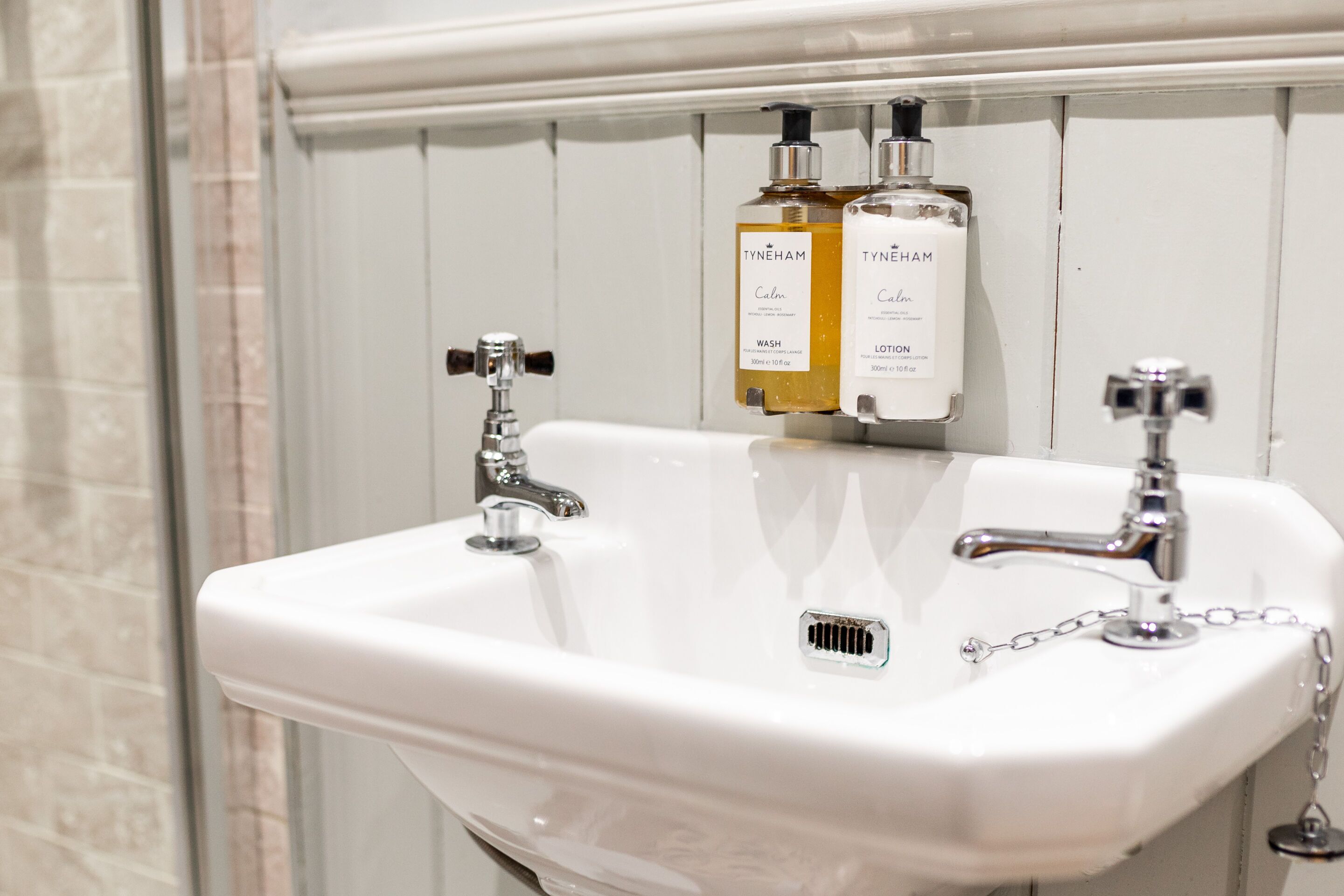 Un lavabo blanco con dos grifos, un espejo y dos botellas de productos TYNEHAM.