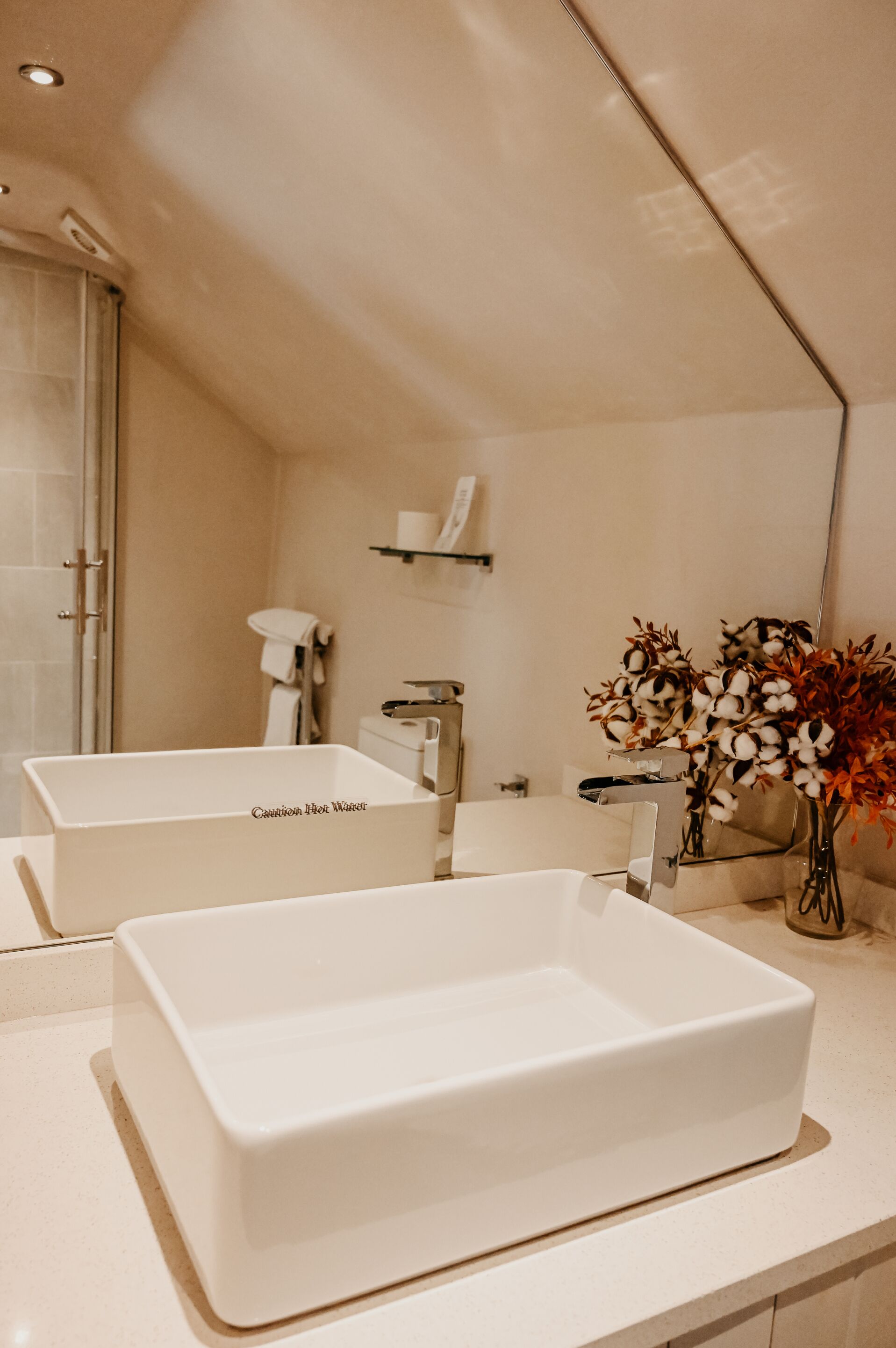 A bathroom with two white sinks, a mirror, a shower, and a vase with flowers.