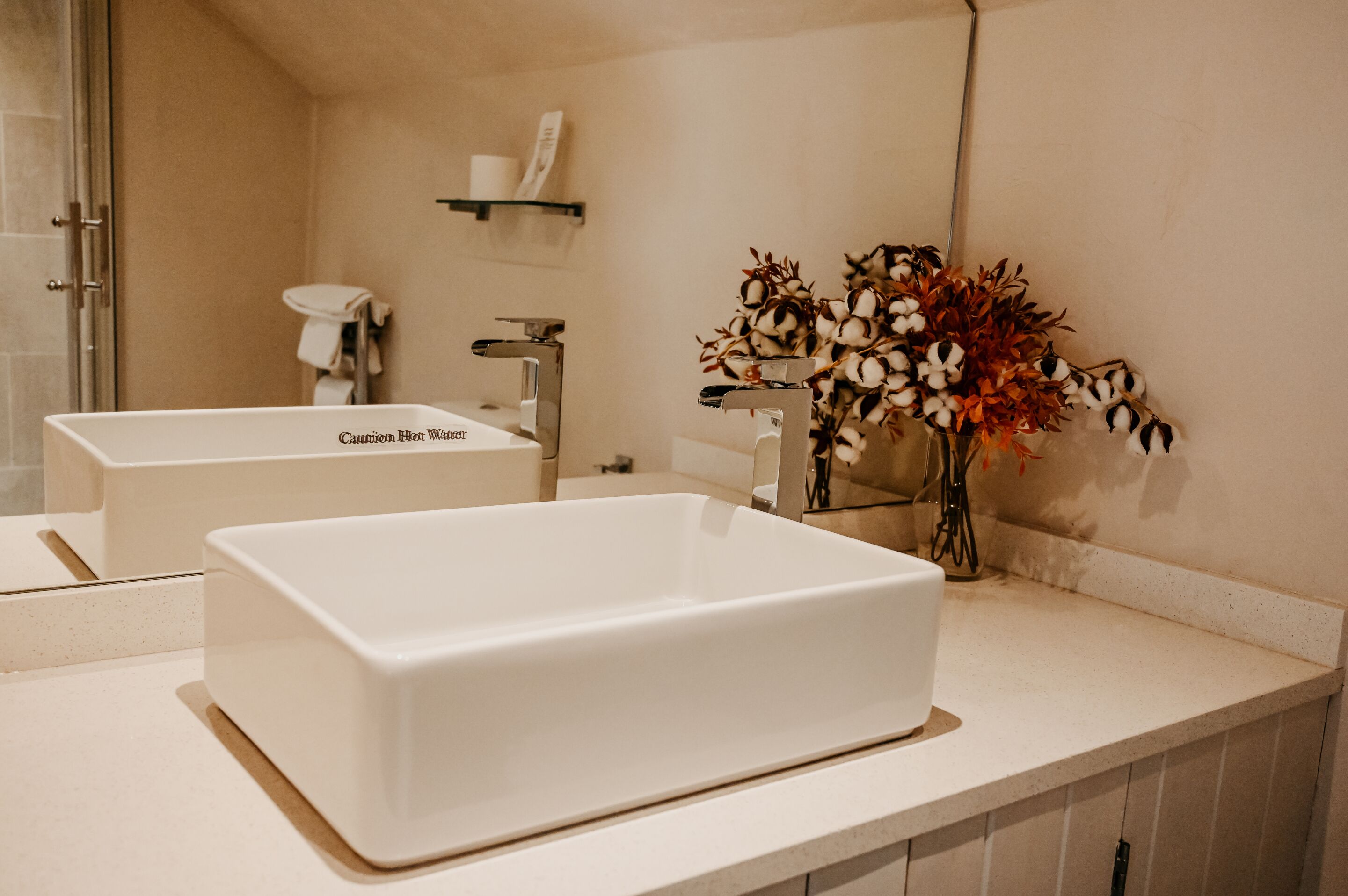 A bathroom with a large white bathtub, a sink, and a mirror. There is a shelf with toiletries and a vase with flowers on the counter.