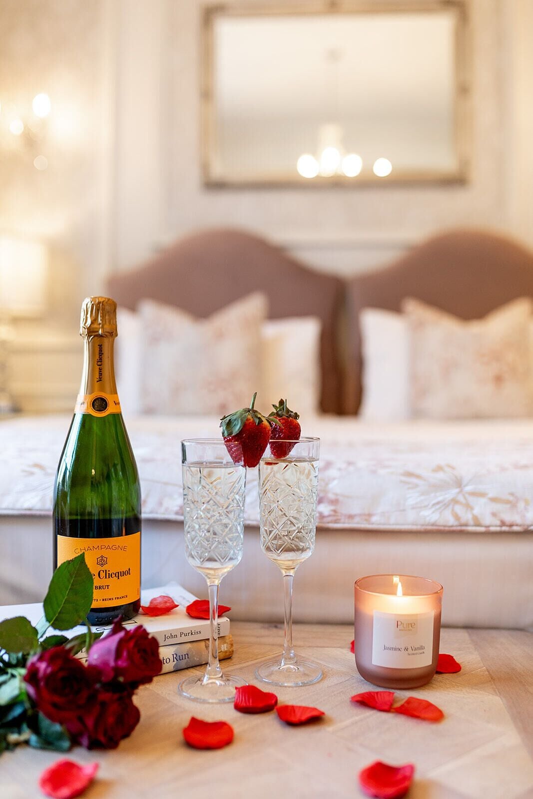Un dormitorio con una cama, una botella de champán, dos copas de champán, una vela encendida y pétalos de rosa esparcidos.