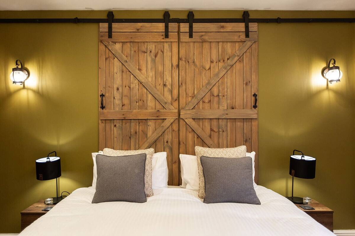 Un dormitorio con cabecera de madera, dos lámparas de noche y una cama con ropa blanca y varias almohadas.