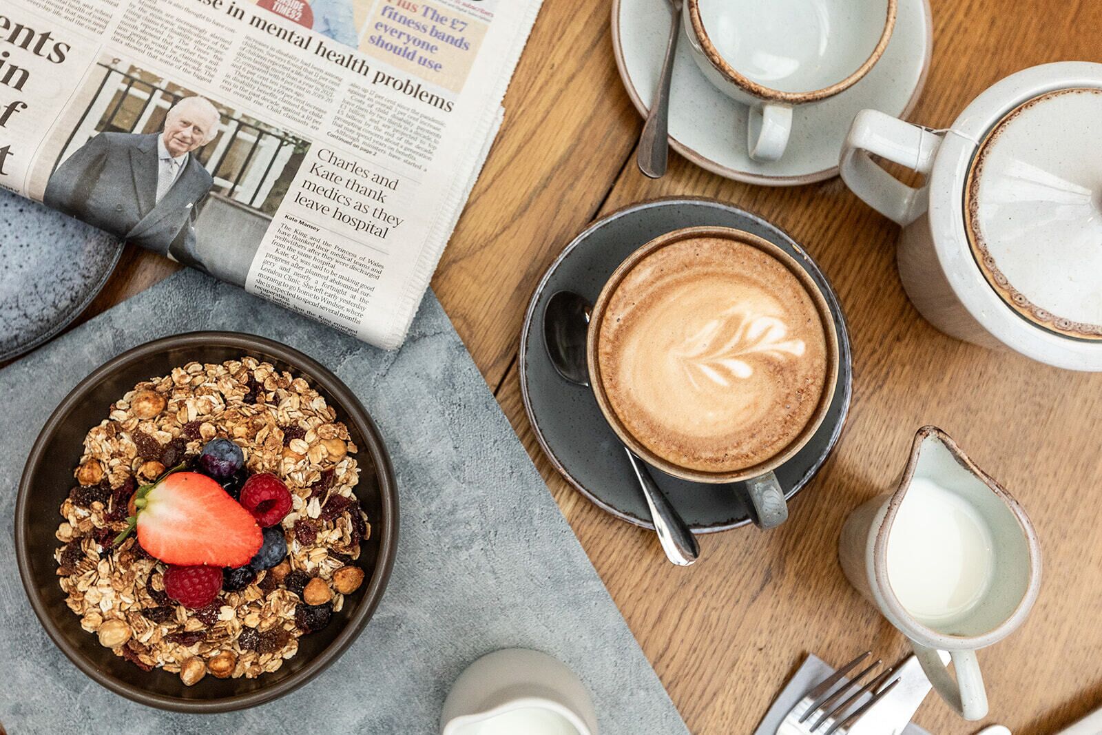 A breakfast setting with a newspaper, a bowl of granola with fruit, a cup of coffee with latte art, and a milk jug.