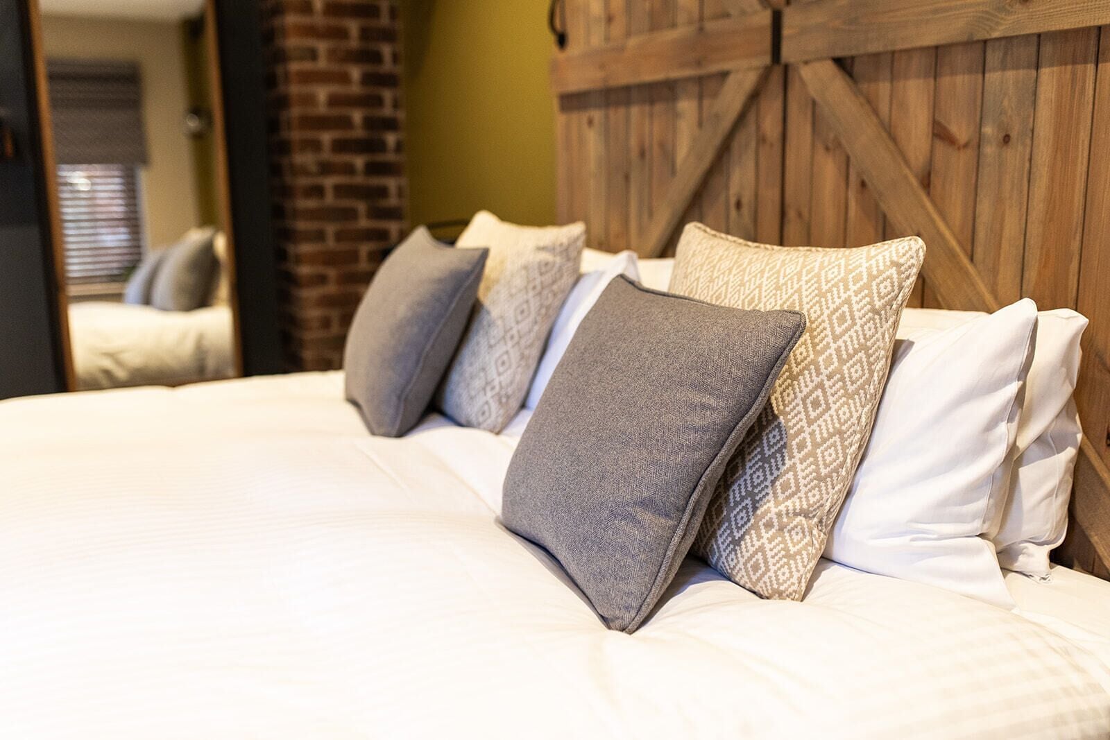 A bed with white bedding and patterned pillows in a room with a wooden headboard and brick wall.
