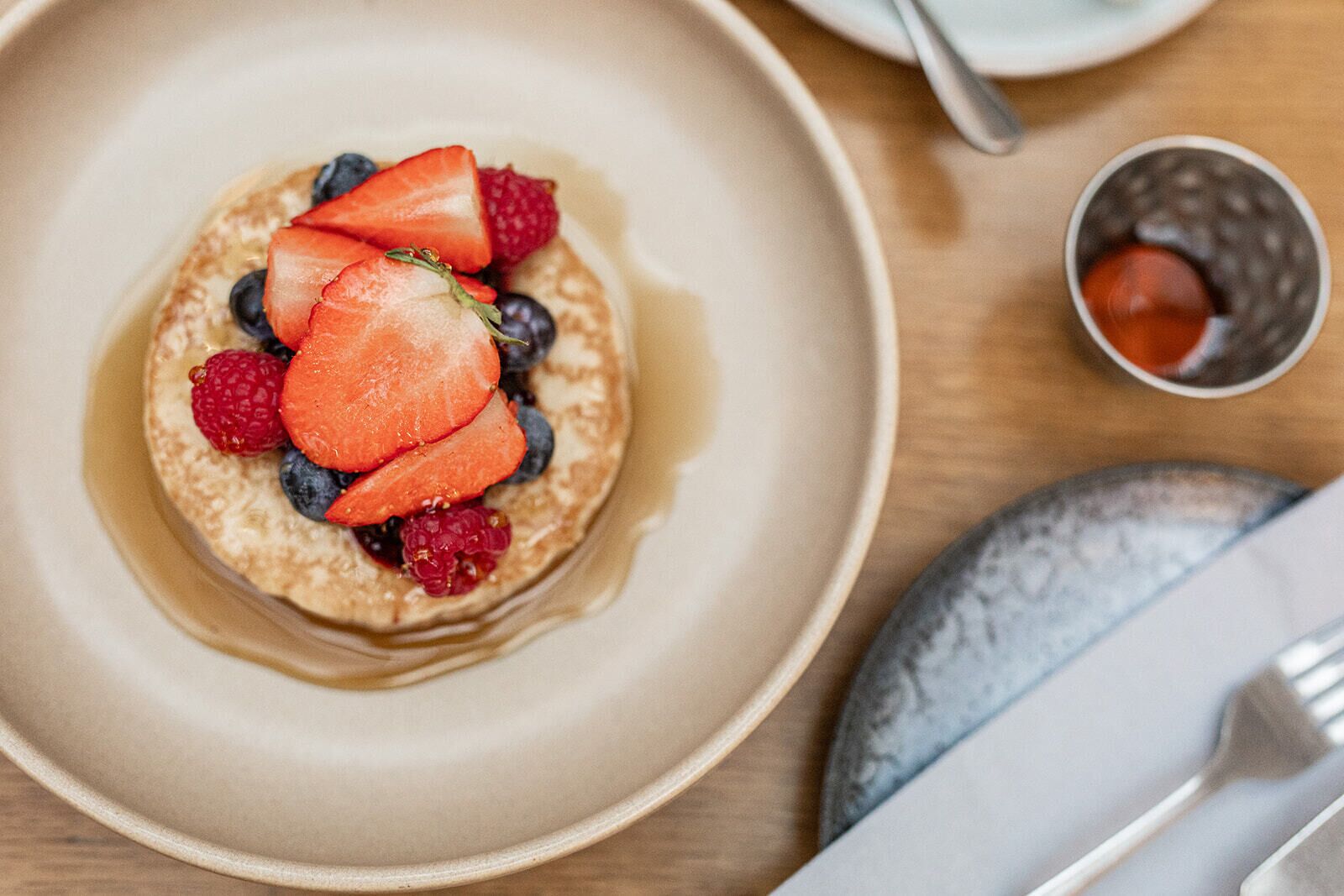 Una porción de panqueques cubiertos de fresas, arándanos y frambuesas, acompañada de almíbar y un tazón pequeño de miel.